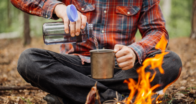 Survival Skills: The 5 Primary Priorities for Survival Man in the forest by a fire, poring some water in his cup. - StayWild Outdoor