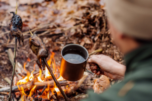 Man by the fire with a cup of coffee, roosting food on the fire. - StayWild Outdoor